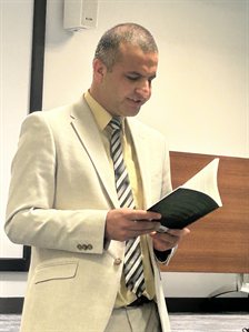 Amir Khesro standing at a lectern, reading aloud from one of his books