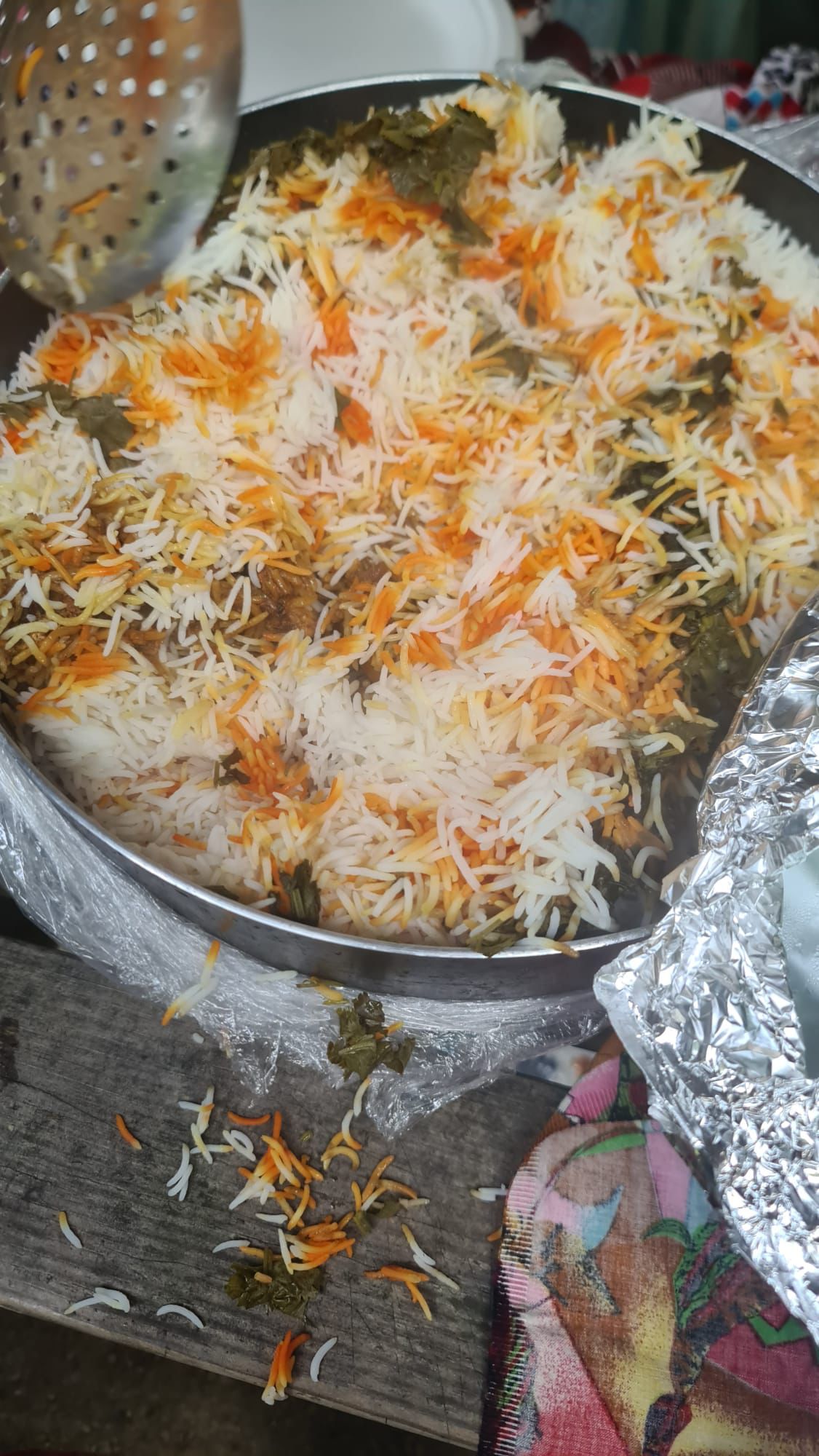 A large metal tray of biryani, the rice streaked orange and white and studded with meat and greens, with a serving spoon resting on the rim