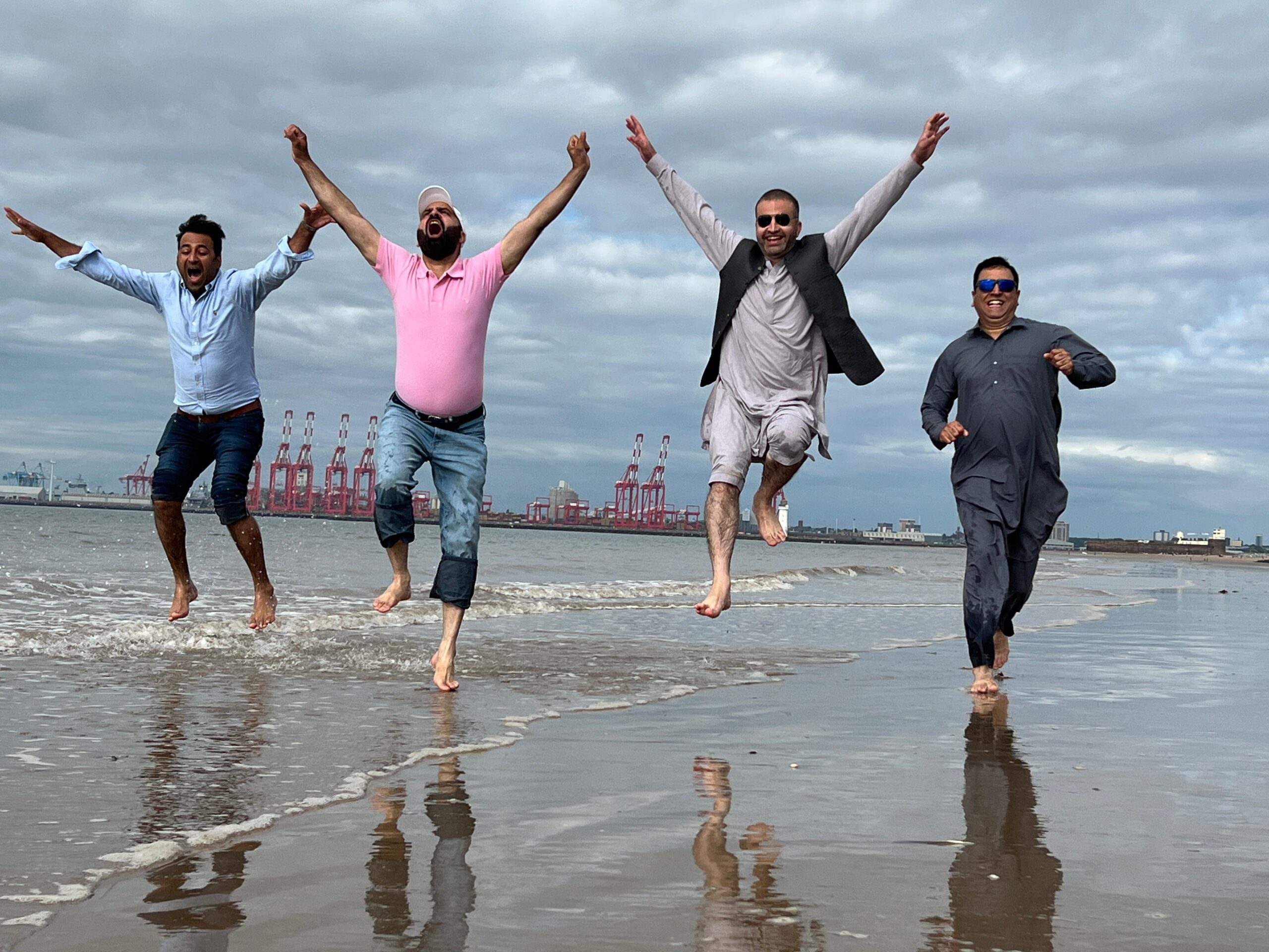 Four men leaping barefoot through the shallow surf with their arms flung wide, the cranes of the Liverpool docks on the horizon behind them