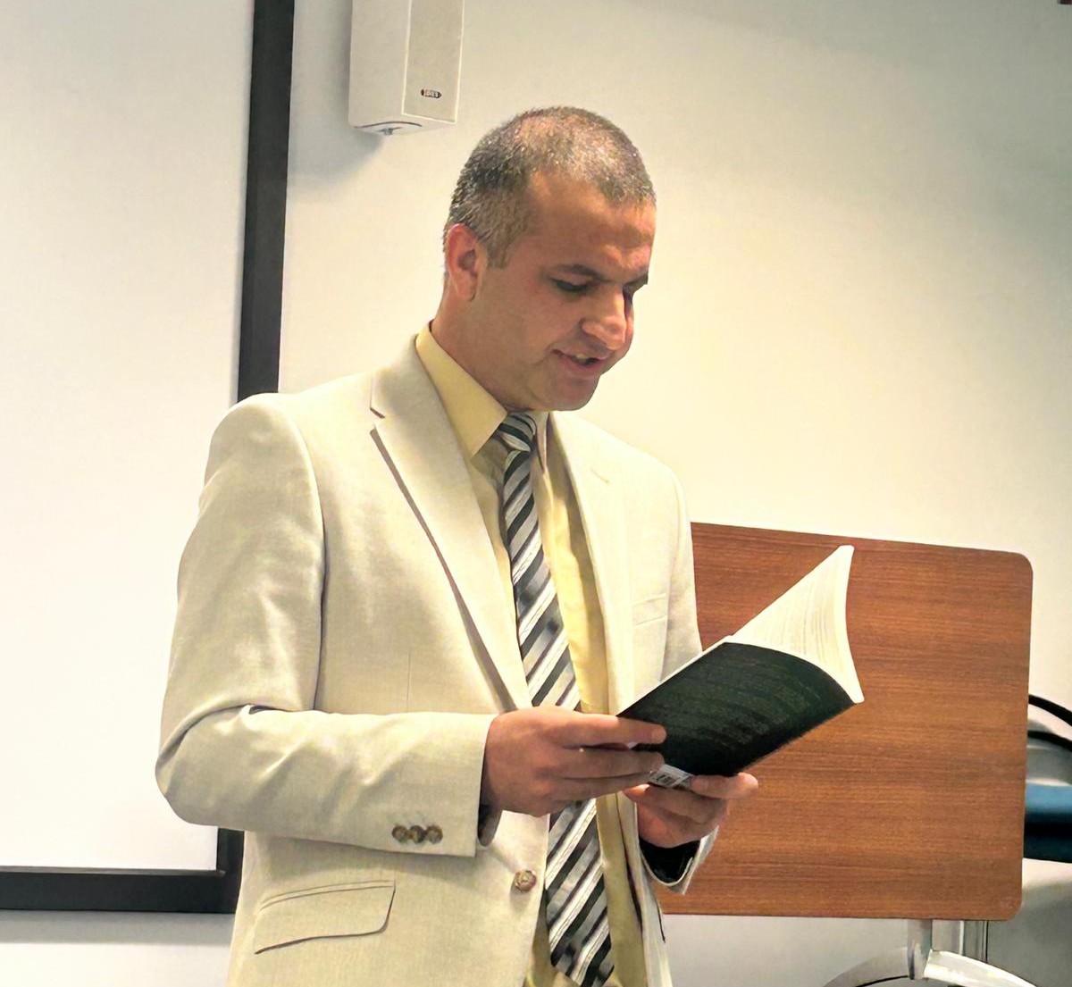 Amir Khesro in a cream suit reading a passage aloud from his book at a lectern in the Salford lecture theatre